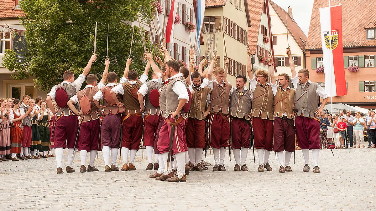Schwerttänzer in roten Pluderhosen und Lederwämsern recken im Kreis ihre Schwerter empor, ringsum Zuschauer