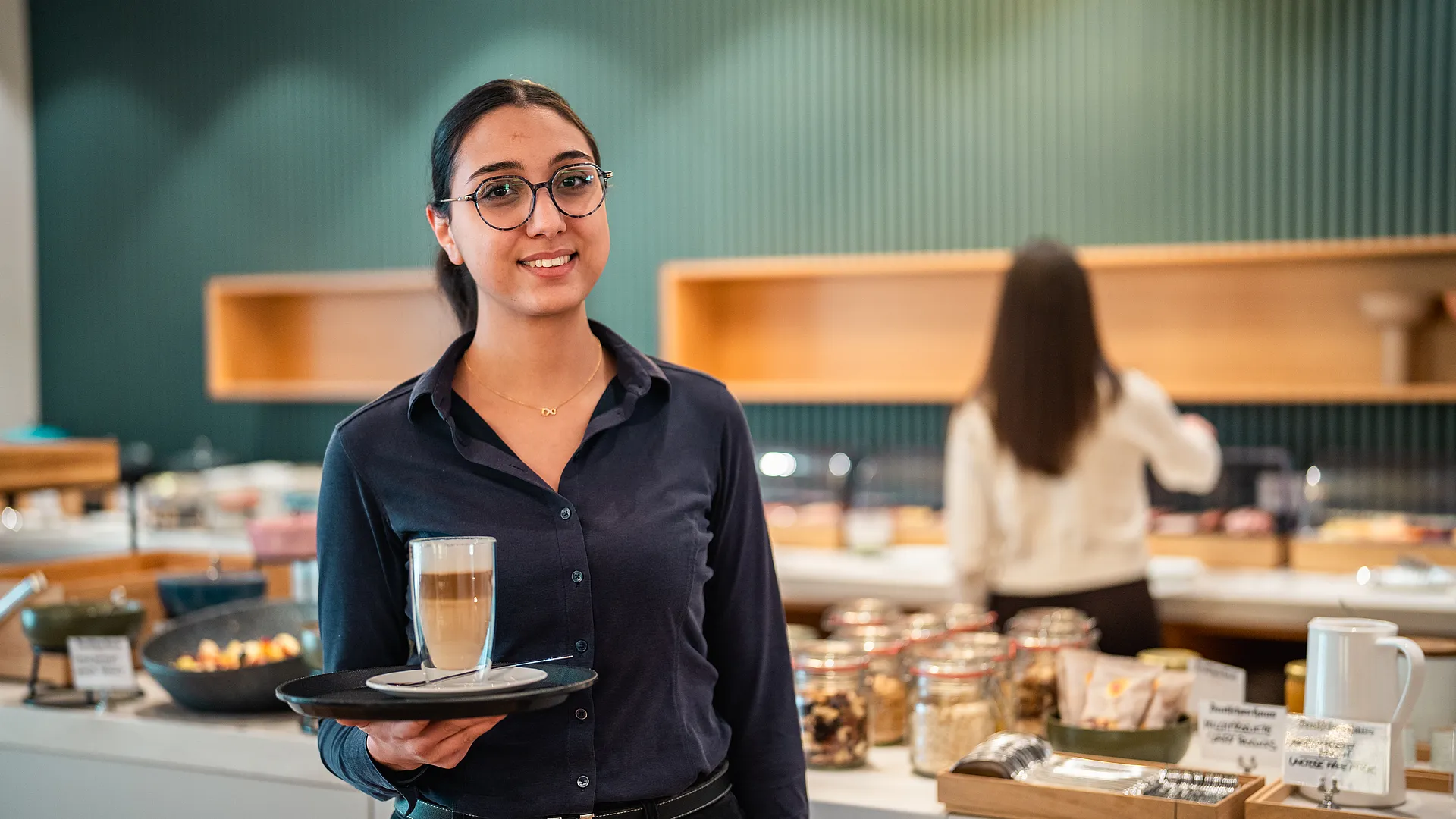 Lächelnde Frau mit Brille trägt Latte macchiato auf einem Tablett, dahinter das Frühstücksbuffet