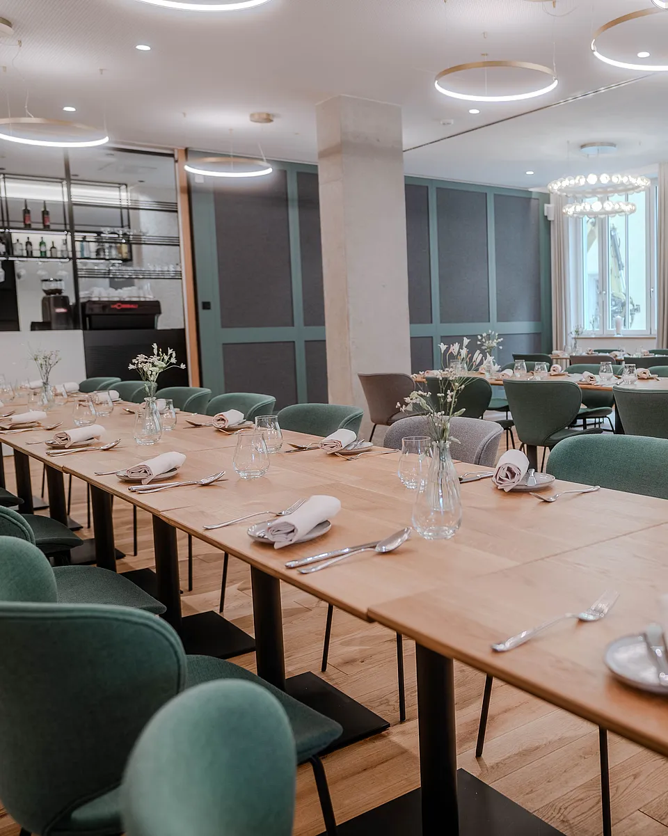 Lange eingedeckte Tafel mit grünen Polsterstühlen, Stoffservietten, Gläsern und Blumen in kleinen Vasen