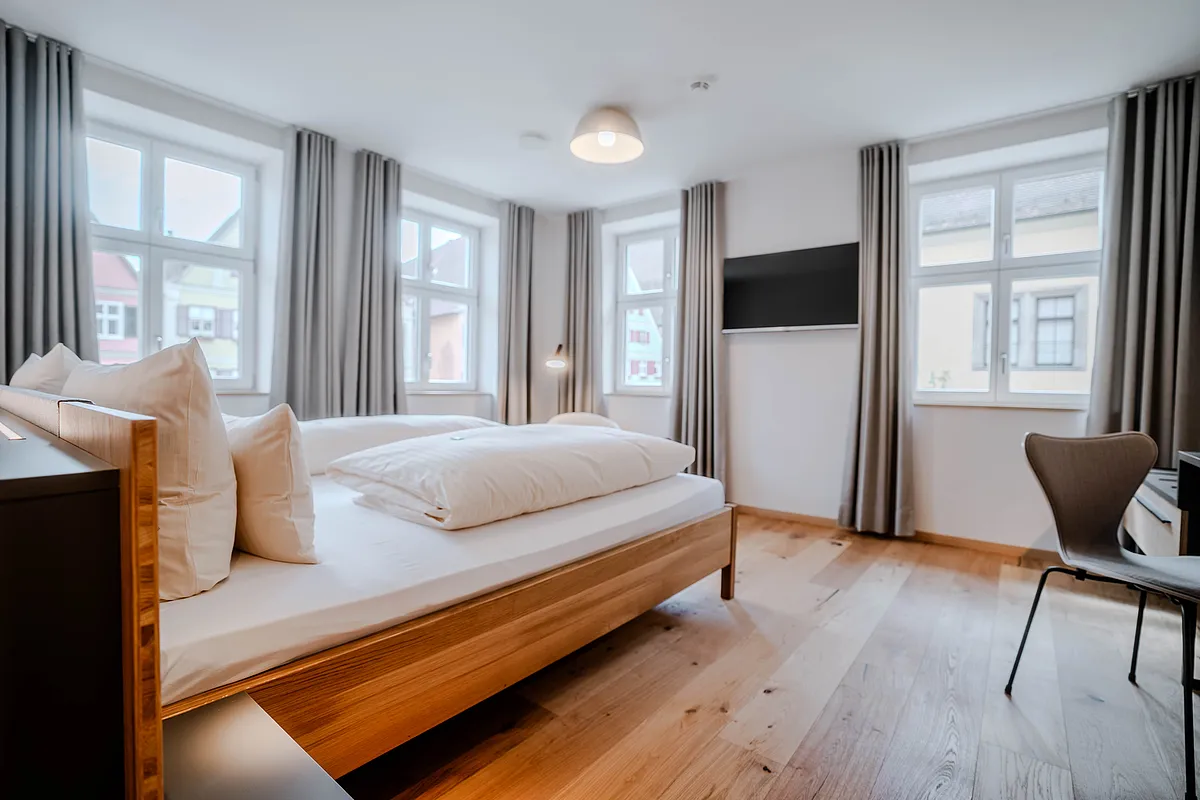 Helles Zimmer mit Holzdoppelbett, Wandfernseher und mehreren Sprossenfenstern mit grauen Vorhängen