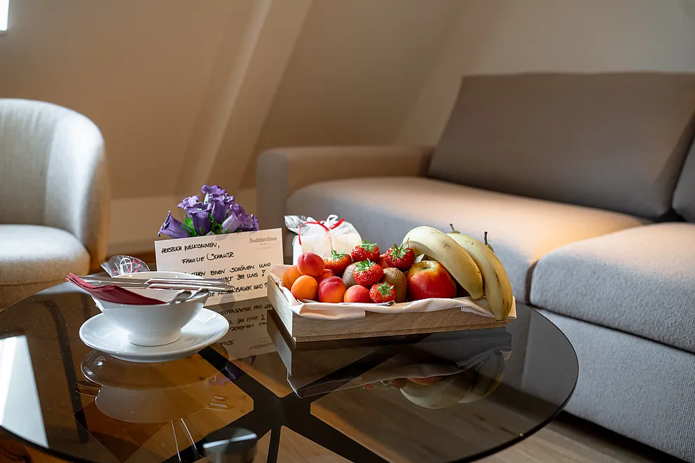 Erdbeeren, Äpfel und Bananen auf einem Holztablett, dazu lila Blumen und eine handgeschriebene Karte auf dem Glastisch der Suite