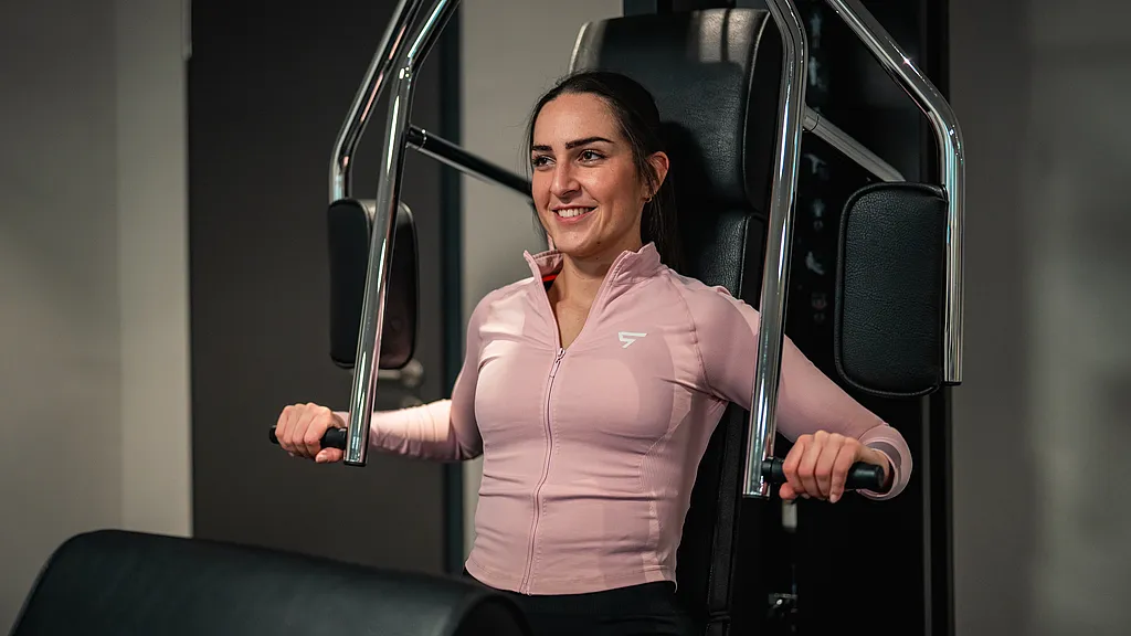 Lächelnde Frau in rosa Sportjacke trainiert an der Brustpresse
