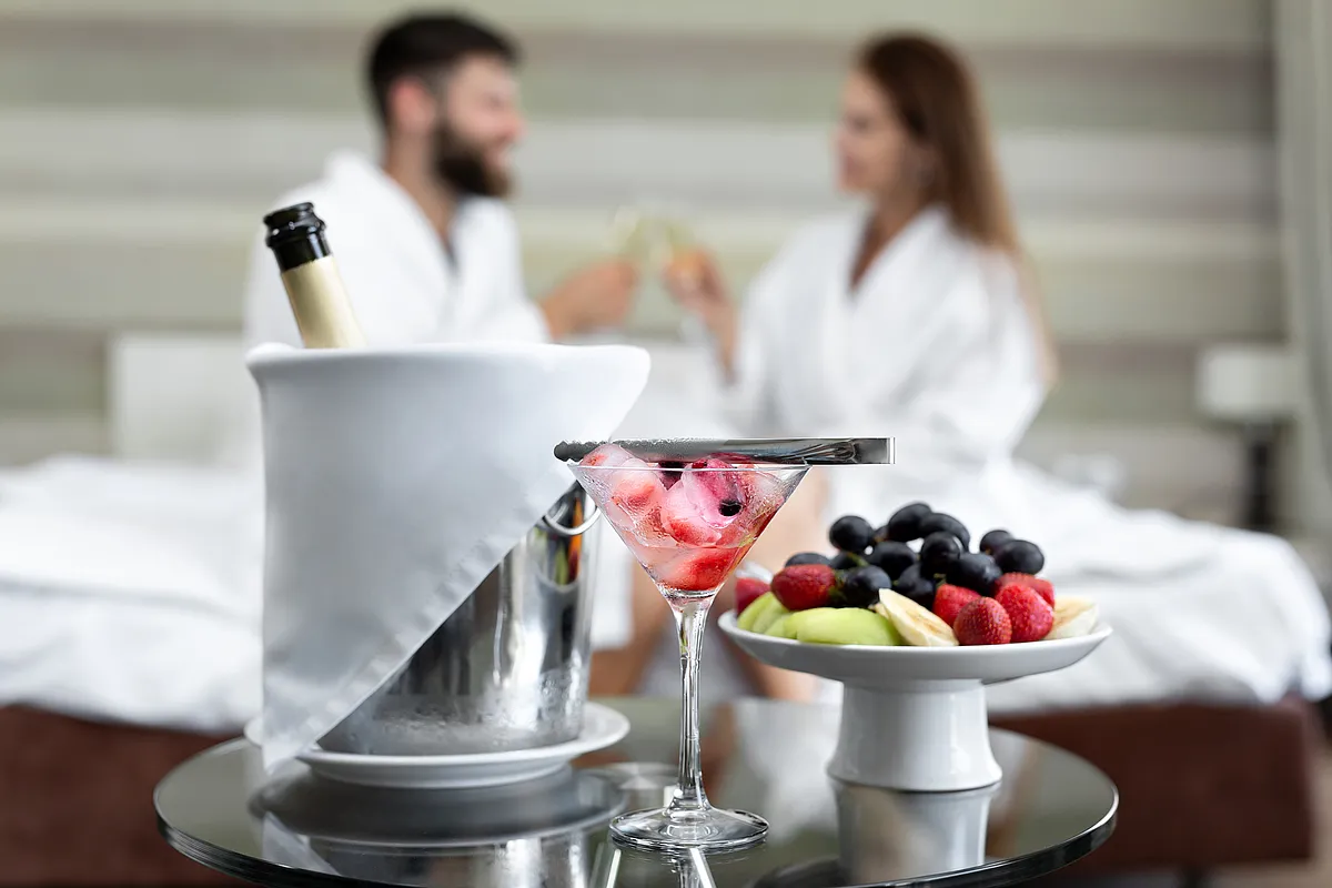 Sektflasche im Kühler, Cocktailglas mit Erdbeeren und Obstschale, dahinter stößt ein Paar in Bademänteln auf dem Bett an