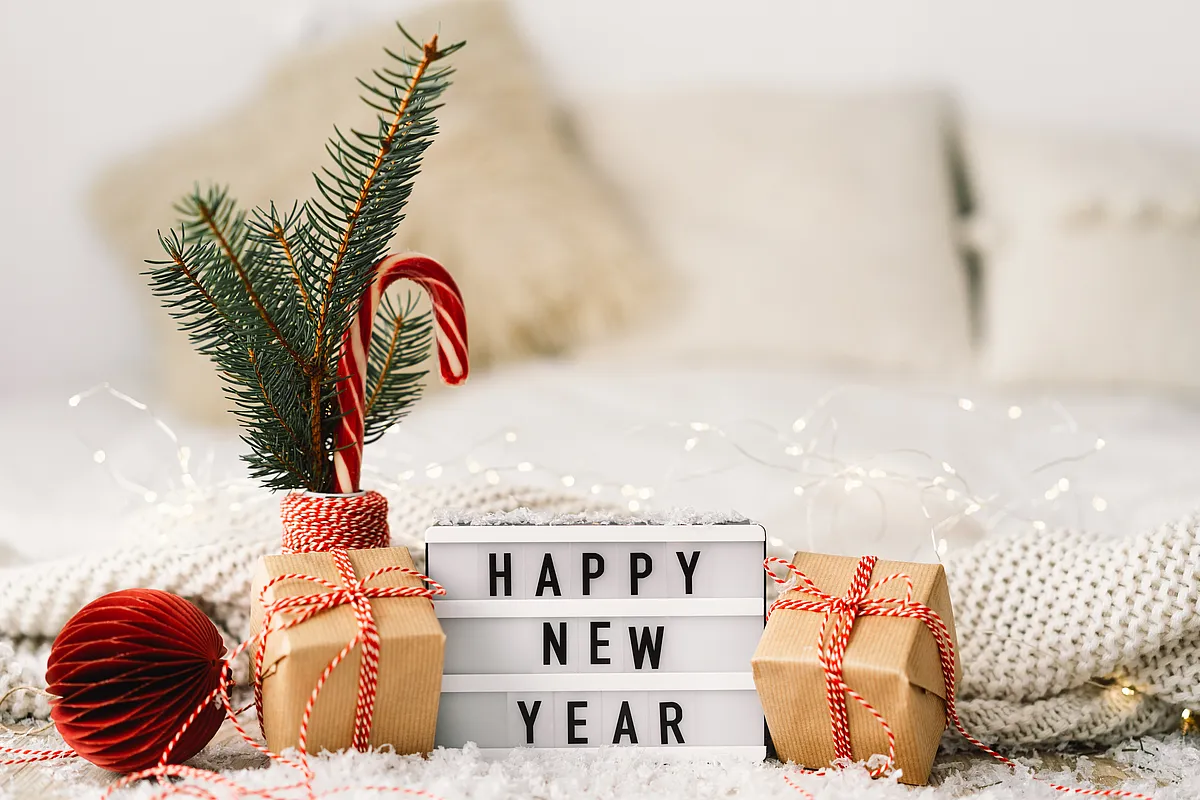 Leuchtkasten mit Schriftzug Happy New Year zwischen Geschenken in Packpapier, Tannenzweig mit Zuckerstange und Lichterkette