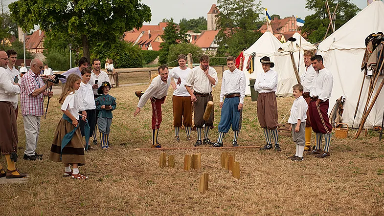 Männer in historischen Gewändern beim Kegelspiel auf einer Wiese, dahinter weiße Zelte und aufgestellte Spieße