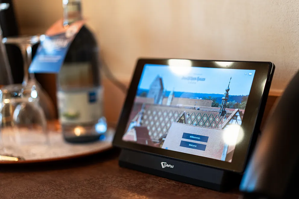 Tablet mit Begrüßungsbildschirm des Hotels in einer Ladestation, im Hintergrund Wasserflasche und Gläser auf einem Serviertablett