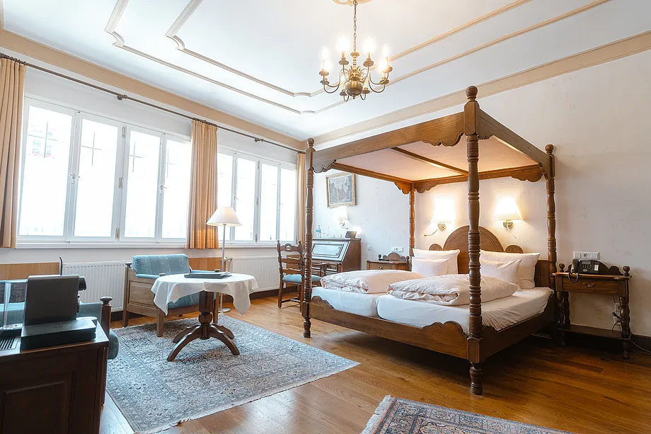 Zimmer mit hölzernem Himmelbett, Kronleuchter, Sitzecke mit rundem Tisch und breiter Fensterfront