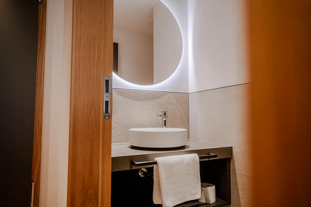 Aufsatzwaschbecken mit rundem Leuchtspiegel im separaten WC der Suite, Blick durch die offene Holztür