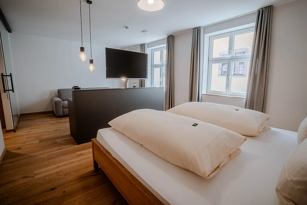 Zimmer mit Holzdoppelbett, Raumteiler mit Fernseher, grauem Sessel und Sprossenfenstern mit grauen Vorhängen