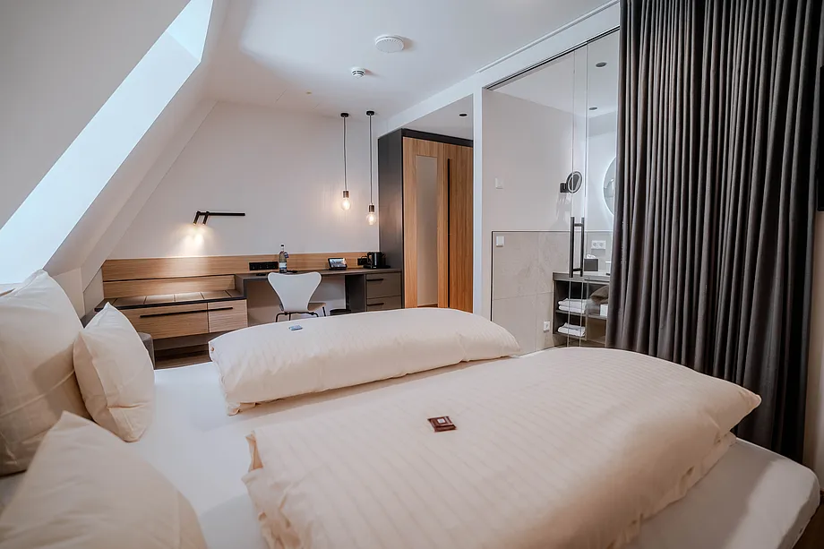 Zimmer unter Dachschräge mit Doppelbett, Schreibtisch, Hängeleuchten und Bad hinter Glaswand mit grauem Vorhang