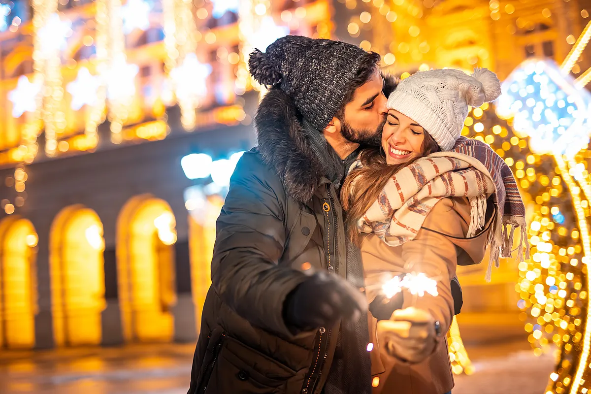 Paar in Wintermützen mit Wunderkerzen vor festlicher Lichterdekoration, der Mann küsst die Frau auf die Wange