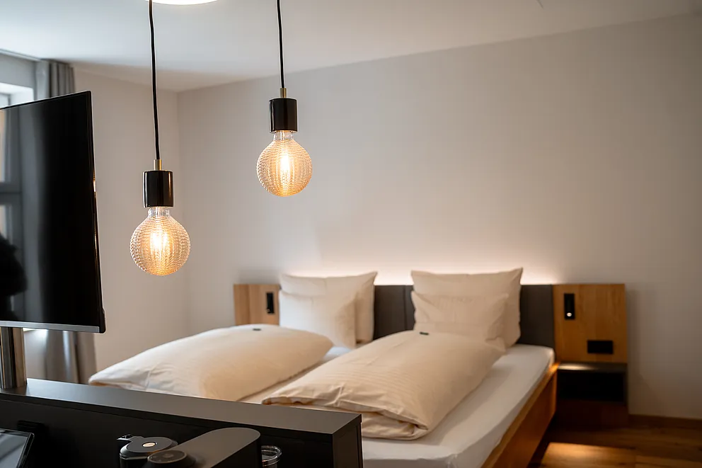 Doppelbett mit grau gepolstertem Kopfteil und Holzpaneelen in der Junior-Suite, davor zwei Pendelleuchten mit Glühbirnen