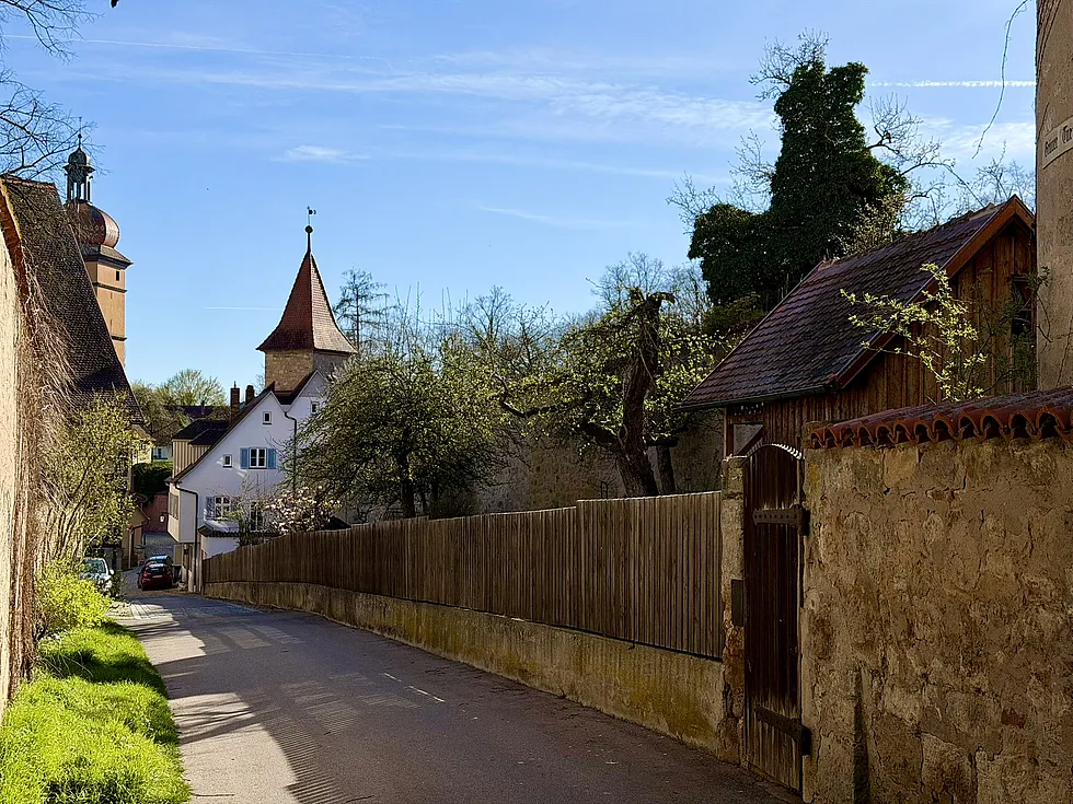 Schmale Straße entlang der Stadtmauer mit Holzzaun und Natursteinmauer, im Hintergrund zwei Türme und ein weißes Haus