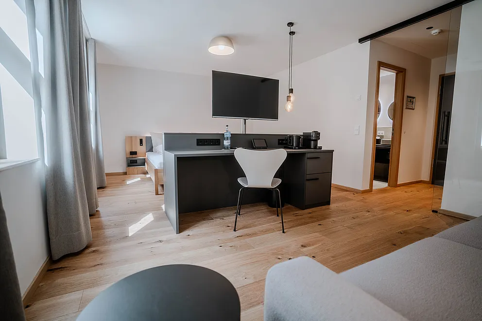 Wohnbereich der Junior-Suite mit Schreibtisch, weißem Stuhl, Wandfernseher, grauem Sofa und Holzdielenboden