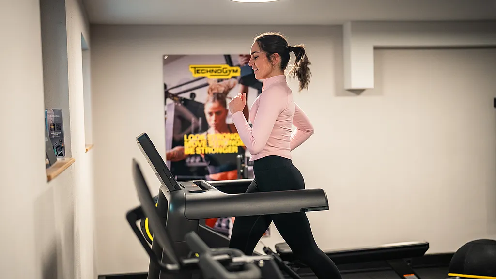 Frau joggt auf dem Laufband, im Hintergrund ein großes Trainingsposter an der Wand