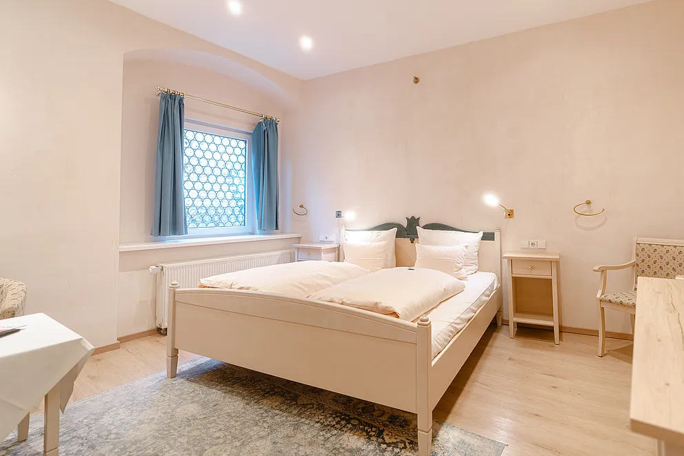 Standard-Doppelzimmer im Stammhaus mit weißem Holzbett, hellem Holzboden und Fenster mit blauen Vorhängen