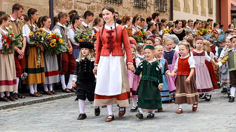 Mädchen in roter Jacke und weißer Schürze führt kleine Kinder in historischen Gewändern an, dahinter Jugendliche mit Blumen