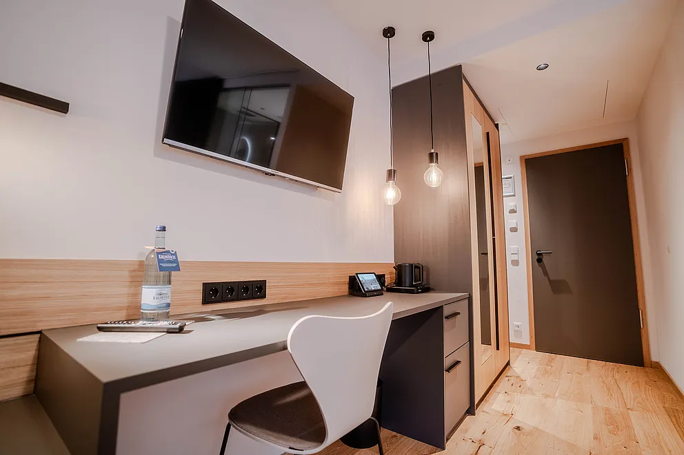Zimmer im Haus Ehemalige Sparkasse mit Schreibtisch, weißem Stuhl, Wandfernseher und zwei Pendelleuchten