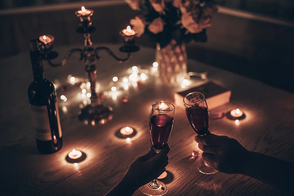 Paar stößt bei Kerzenschein mit Rotwein an, auf dem Tisch Kerzenleuchter, Teelichter, Lichterkette und Rosen