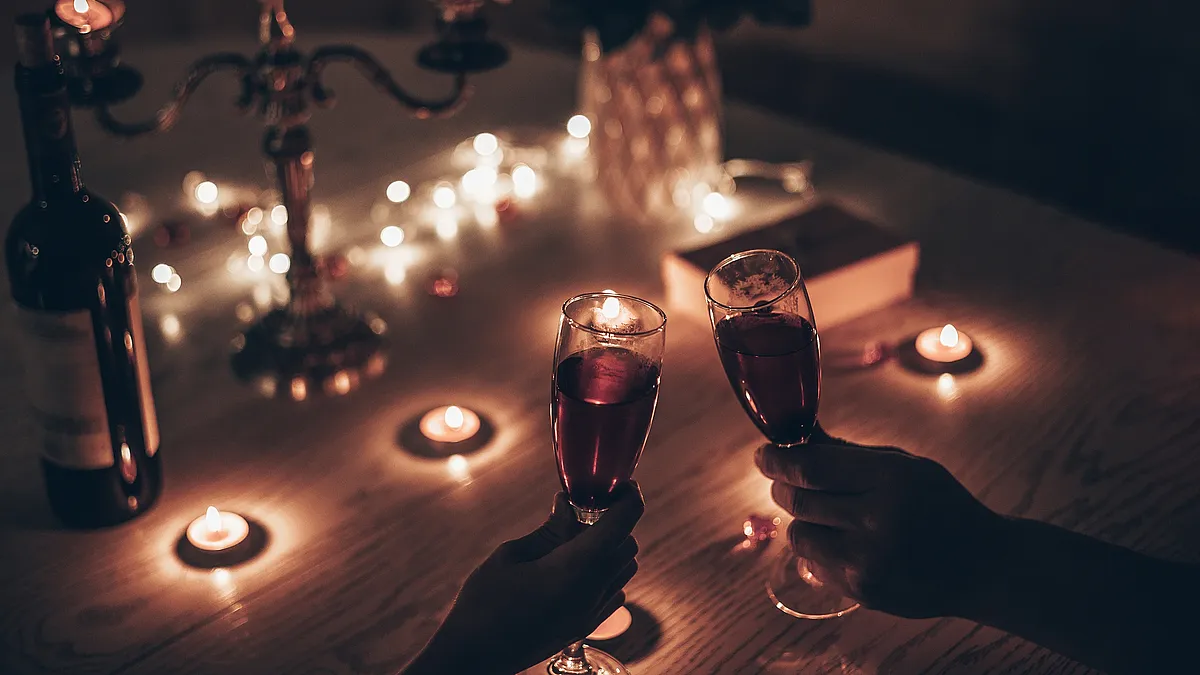 Paar stößt bei Kerzenschein mit Rotwein an, auf dem Tisch Kerzenleuchter, Teelichter, Lichterkette und Rosen