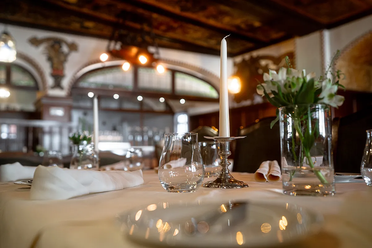 Eingedeckter Tisch mit weißer Kerze in silbernem Leuchter, Gläsern und weißen Blumen, dahinter der Gastraum mit bemalter Holzdecke
