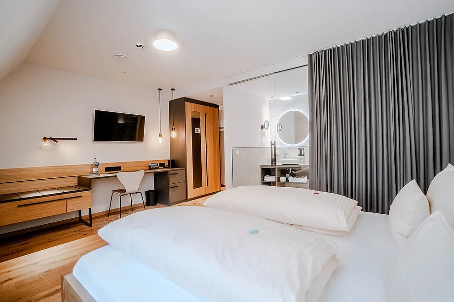 Zimmer unter Dachschräge mit Doppelbett, Schreibtisch, Wandfernseher und Bad hinter Glaswand mit rundem Spiegel