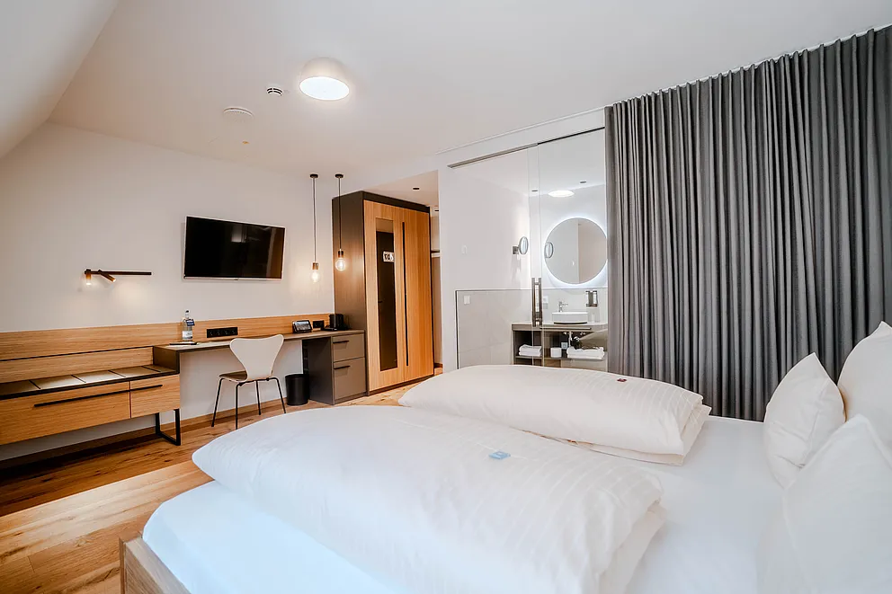 Zimmer unter Dachschräge mit Doppelbett, Schreibtisch, Wandfernseher und Bad hinter Glaswand mit rundem Spiegel