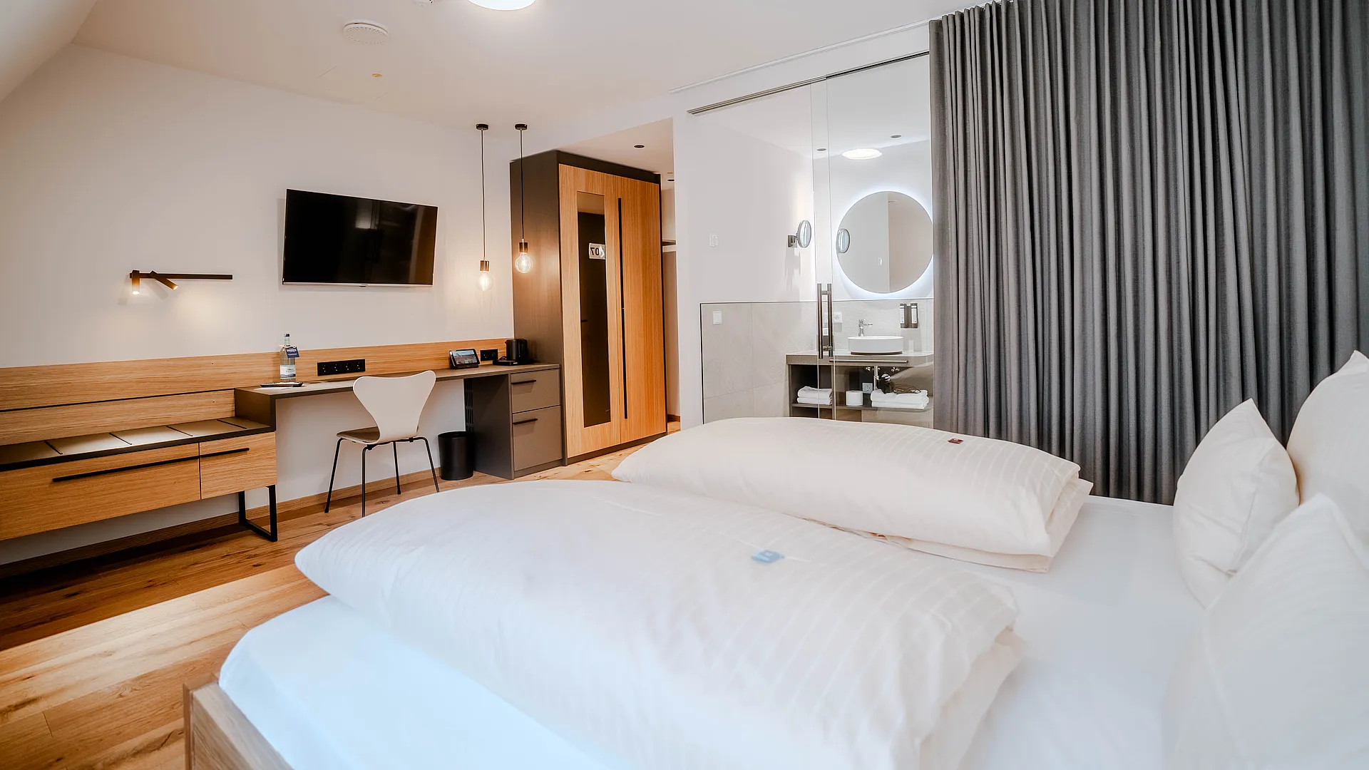 Zimmer unter Dachschräge mit Doppelbett, Schreibtisch, Wandfernseher und Bad hinter Glaswand mit rundem Spiegel