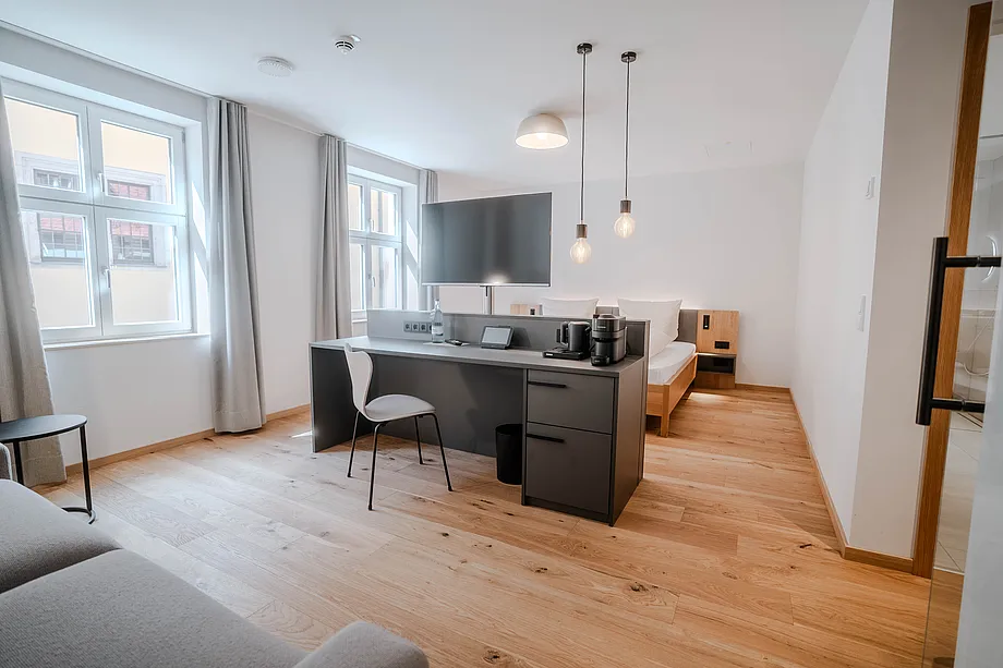 Helles Zimmer mit Sprossenfenstern, langem Schreibtisch mit Fernseher, Bett im Hintergrund und grauem Sofa vorne