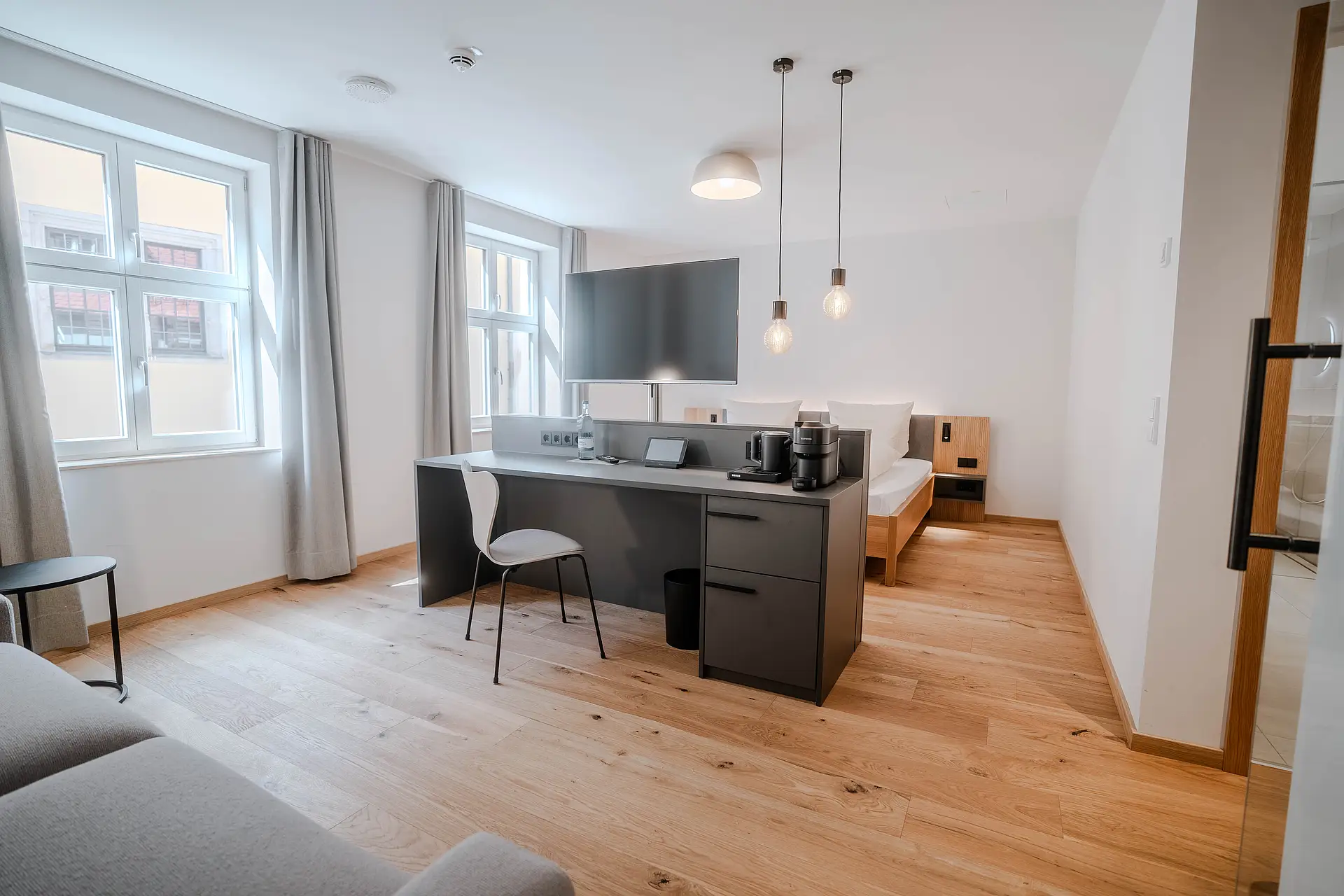 Helles Zimmer mit Sprossenfenstern, langem Schreibtisch mit Fernseher, Bett im Hintergrund und grauem Sofa vorne