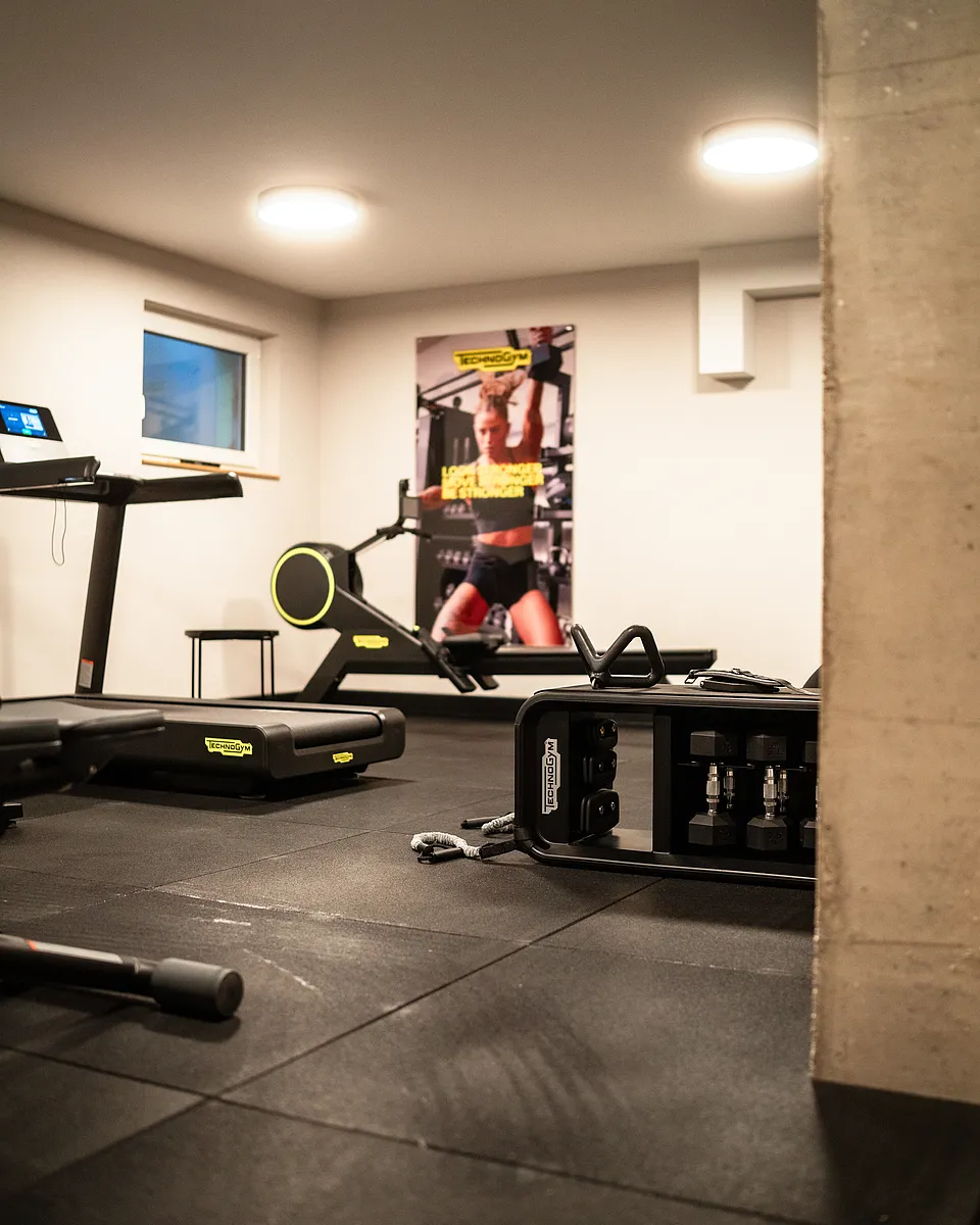 Blick in den Fitnessraum mit schwarzem Gummiboden, Laufband, Rudergerät und Kurzhanteln neben einer Betonsäule