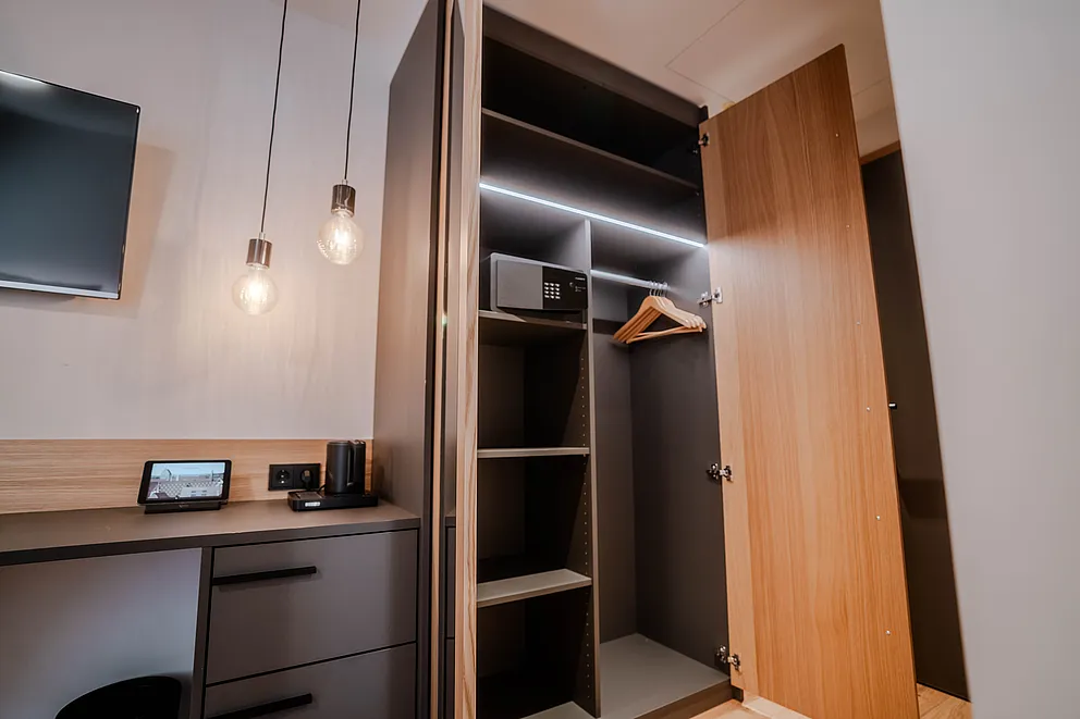 Offener Kleiderschrank mit Safe und Kleiderbügel neben Schreibtisch und Wandfernseher im Komfortzimmer