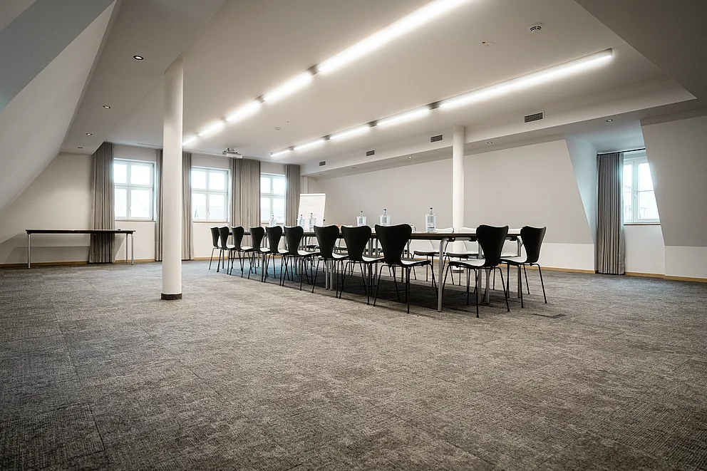Großer Tagungsraum unter Dachschrägen mit langer Tischreihe, schwarzen Stühlen und Lichtbändern an der Decke