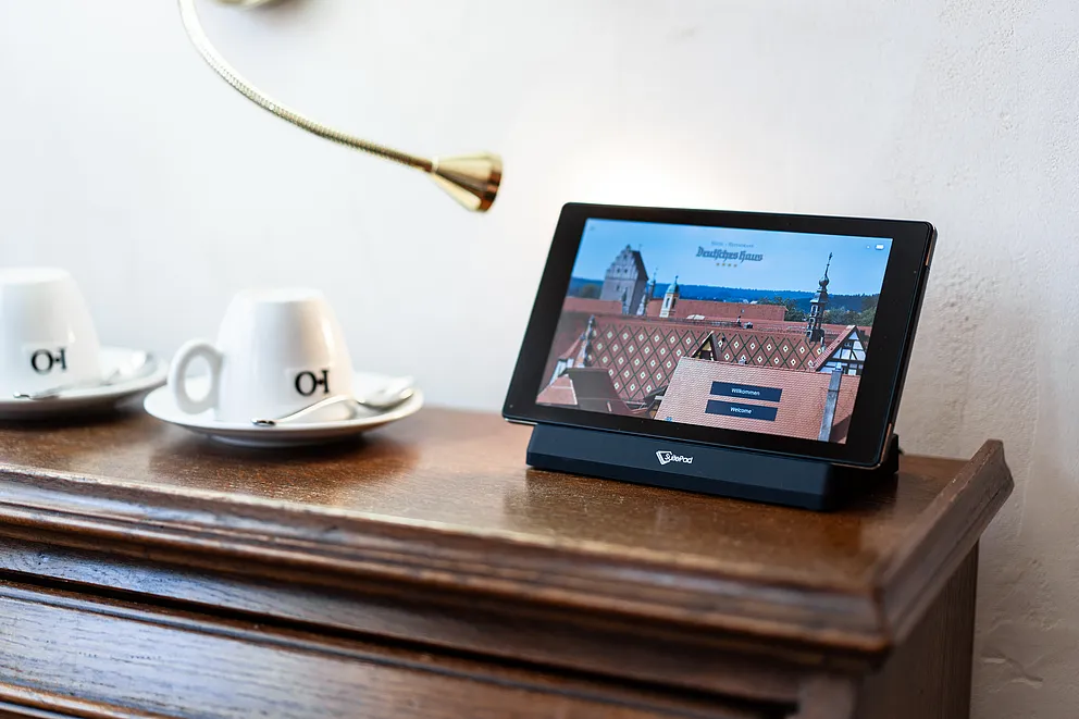 Tablet mit Begrüßungsbildschirm des Hotels auf einer Holzkommode in der Hochzeitssuite, daneben zwei weiße Kaffeetassen