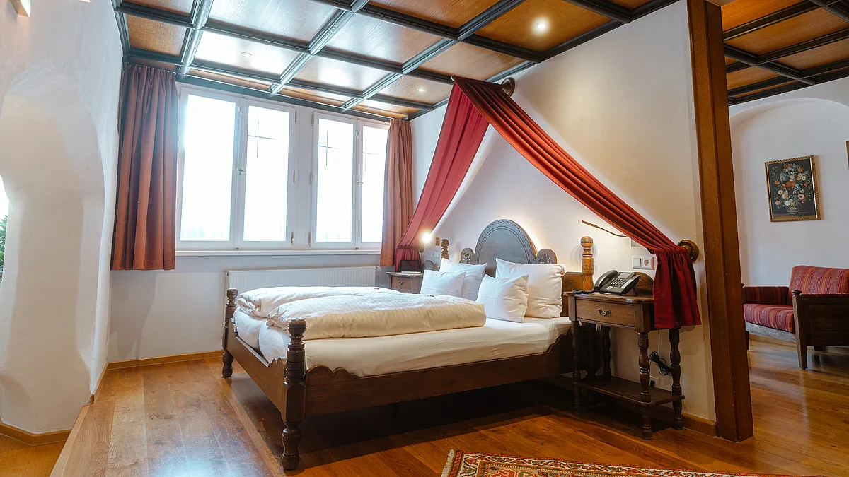 Zimmer mit Kassettendecke aus Holz, Doppelbett unter rotem Stoffbaldachin, Sprossenfenstern und rotem Sofa