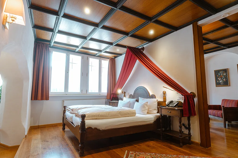 Zimmer mit Kassettendecke aus Holz, Doppelbett unter rotem Stoffbaldachin, Sprossenfenstern und rotem Sofa