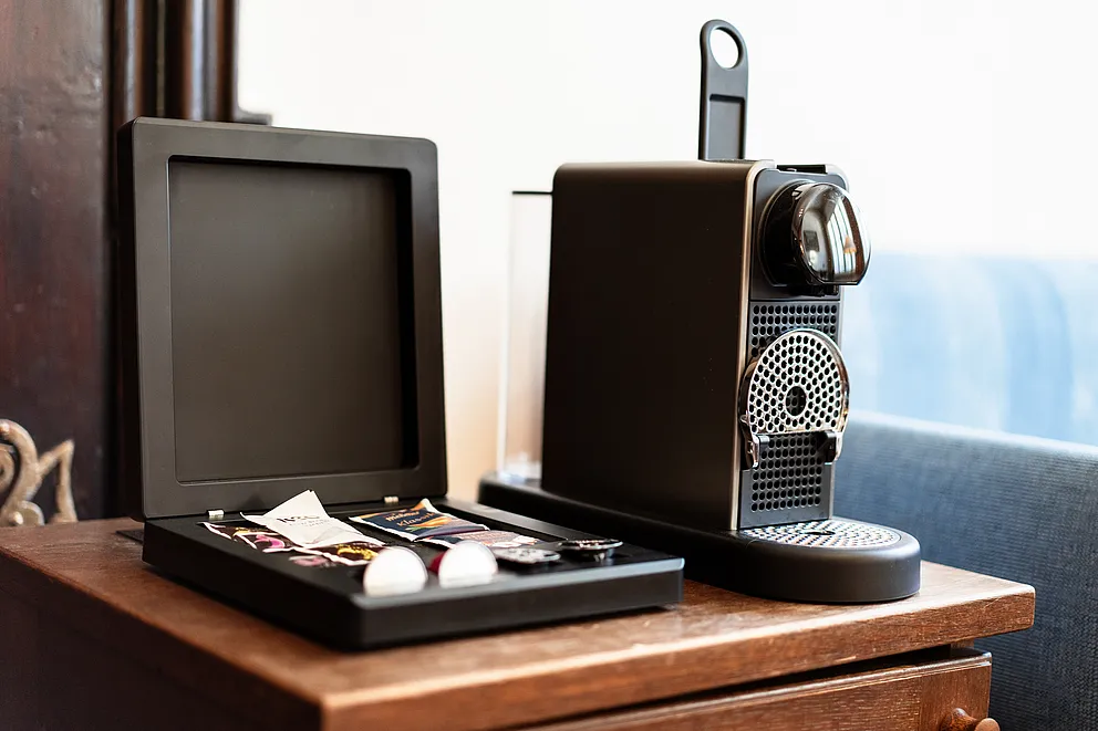 Kapselkaffeemaschine und offene schwarze Box mit Kaffeekapseln auf einer Holzkommode in der Hochzeitssuite im Stammhaus