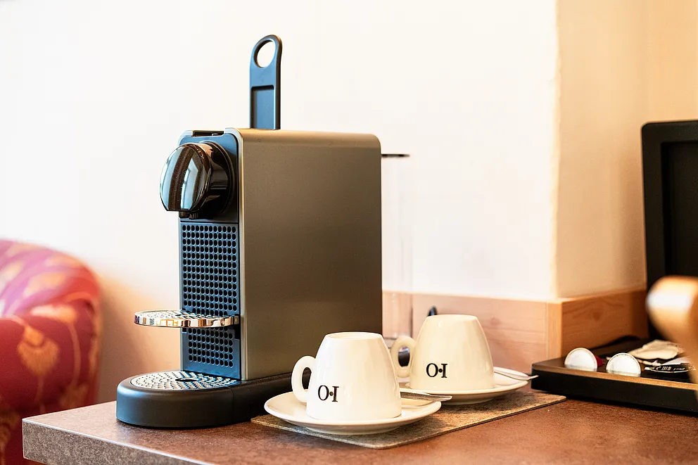 Kapselkaffeemaschine und zwei weiße Tassen auf Untertassen im Superior-Zimmer, links ein rot gemusterter Polstersessel