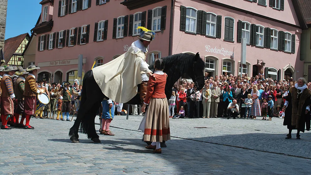 Reiter mit Federhut und weißem Umhang auf schwarzem Pferd, neben ihm ein Mädchen in historischem Gewand, dahinter Zuschauer