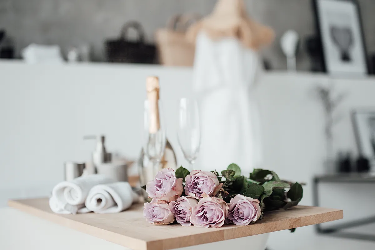 Zartrosa Rosen, gerollte Handtücher, Sektflasche und zwei Gläser auf einem Holzbrett