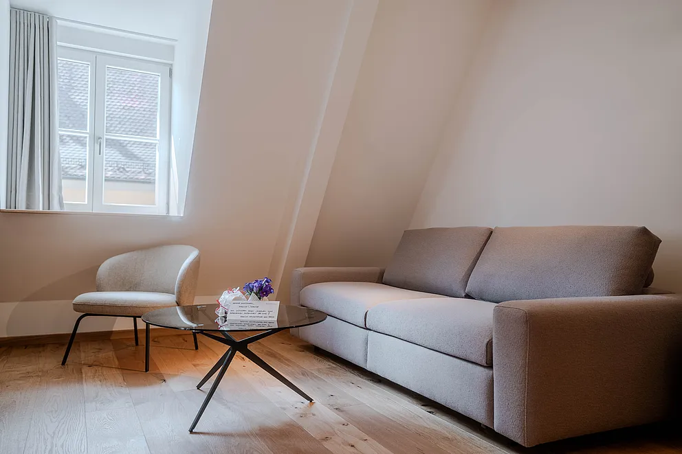 Sitzecke der Suite im Haus Ehemalige Sparkasse mit grauem Sofa, Sessel und rundem Glastisch unter der Dachschräge
