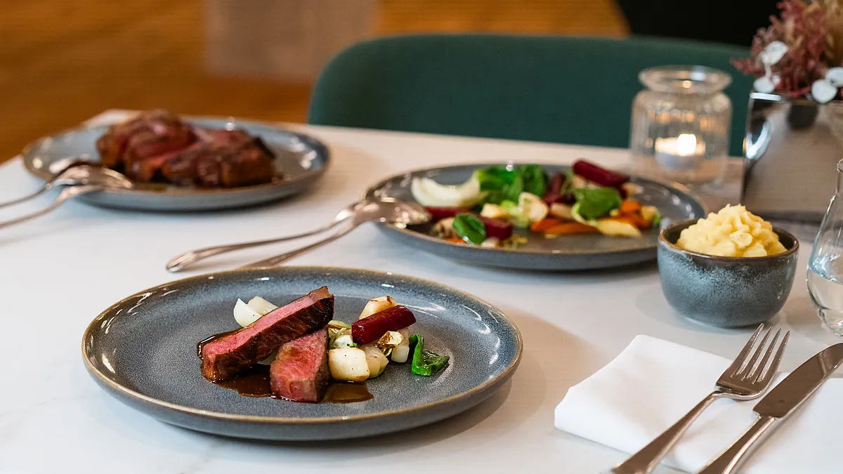 Rosa gebratenes Steak in Scheiben mit Jus und Gemüse auf grauem Teller, dahinter weiteres Fleisch, Gemüse und Kartoffelpüree