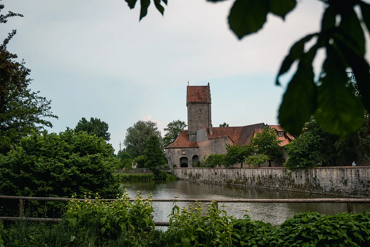 Blick über das Wasser auf einen hohen Turm mit Ziegeldach und ein Gebäude mit Rundbögen hinter einer Ufermauer