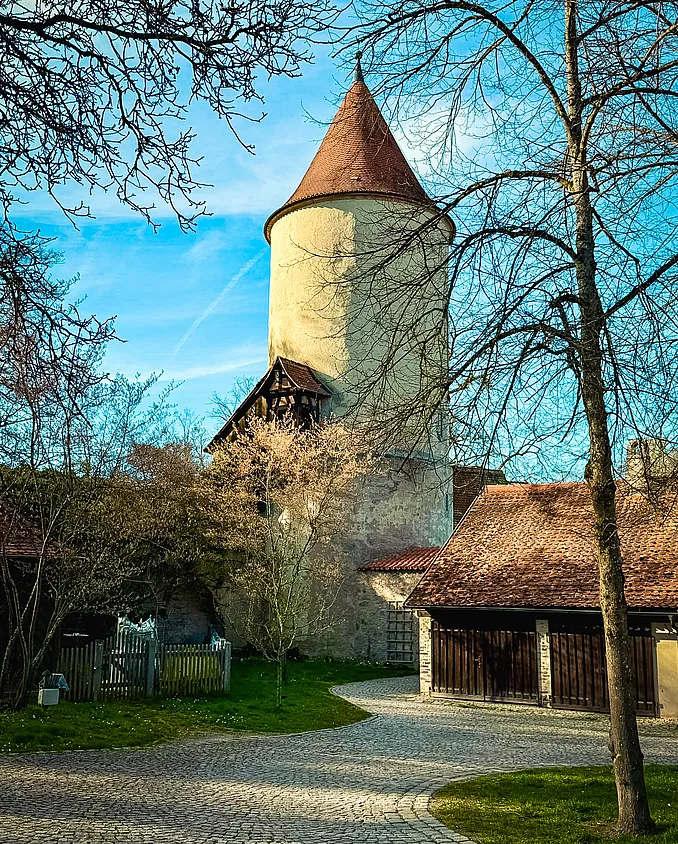 Runder Turm mit Kegeldach und angebautem Fachwerkgiebel, davor kahle Bäume, ein gepflasterter Weg und Schuppen mit Holztoren