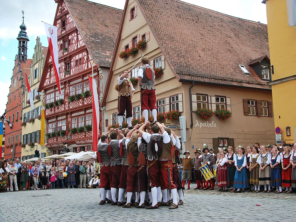 Schwerttänzer in historischer Tracht heben zwei Mittänzer auf ihren Schwertern empor, dahinter Fachwerkhäuser und Zuschauer