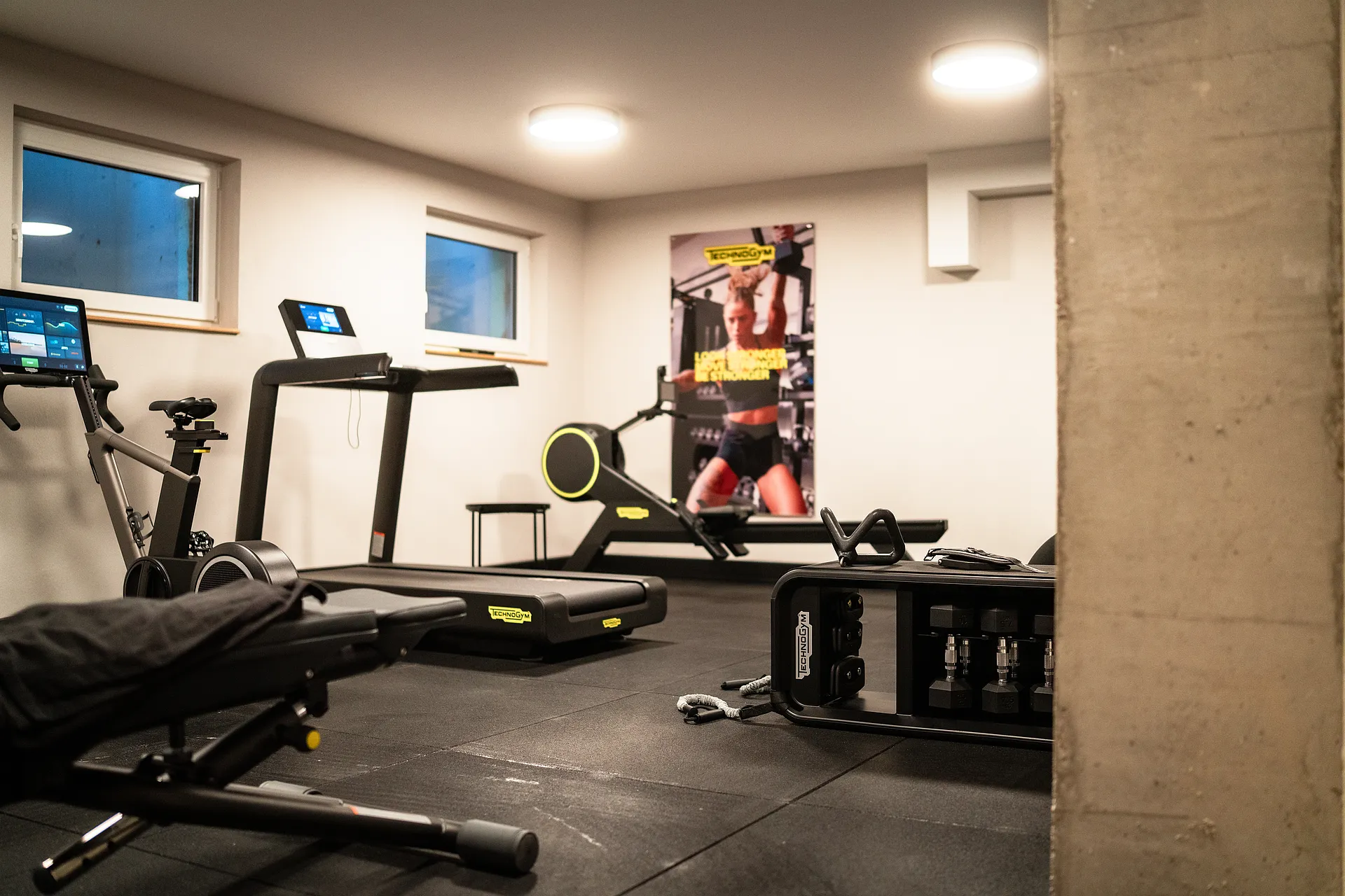 Laufband, Ergometer, Rudergerät und Kurzhantelständer im Fitnessraum, an der Wand ein großes Trainingsposter