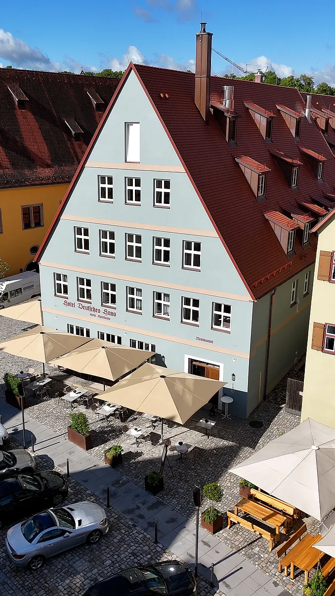 Luftaufnahme des hellblauen Hotelhauses Ehemalige Sparkasse mit Ziegeldach und Sonnenschirmen auf dem Platz davor
