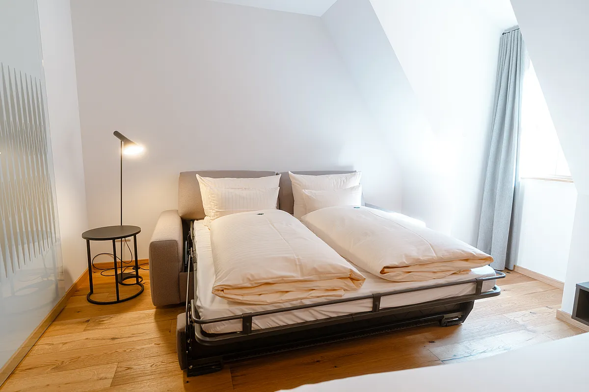 Ausgezogenes Schlafsofa mit weißer Bettwäsche für zwei Personen unter der Dachschräge, daneben Stehlampe und Beistelltisch