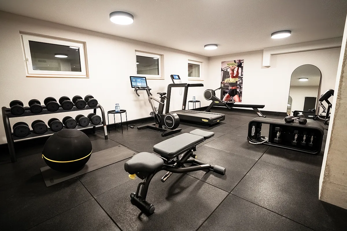 Fitnessraum mit Hantelablage, Laufband, Ergometer, Hantelbank und Gymnastikball auf schwarzem Gummiboden