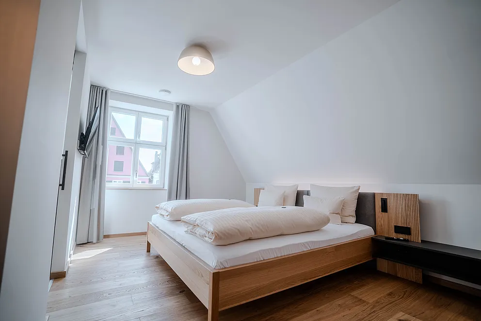 Schlafzimmer im Haus Ehemalige Sparkasse mit Doppelbett unter der Dachschräge, Pendelleuchte und Fenster mit grauen Vorhängen